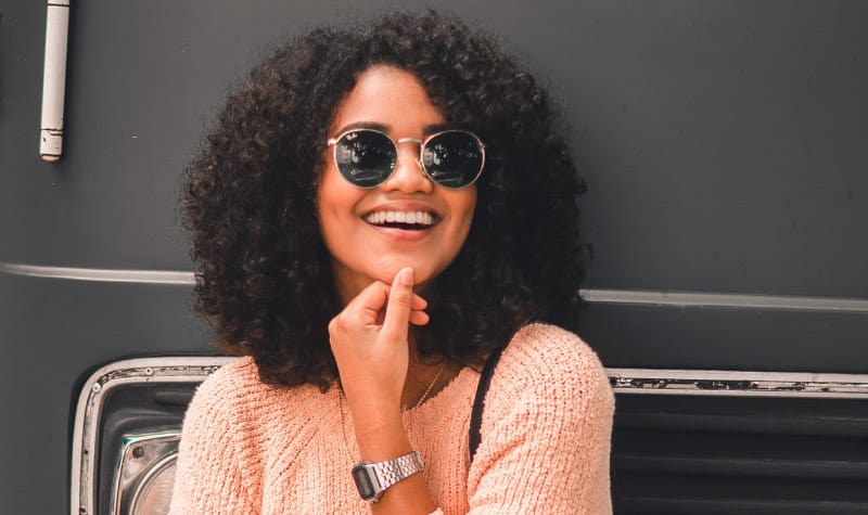 Curly-haired woman wearing sunglasses, a peach sweater, and a silver watch smiles after BOTOX relieved her facial pain